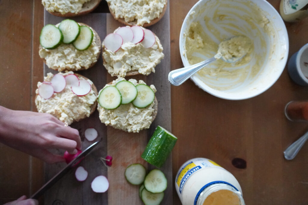 woman slicing radishes on a cutting board beside open-faced egg salad bagels