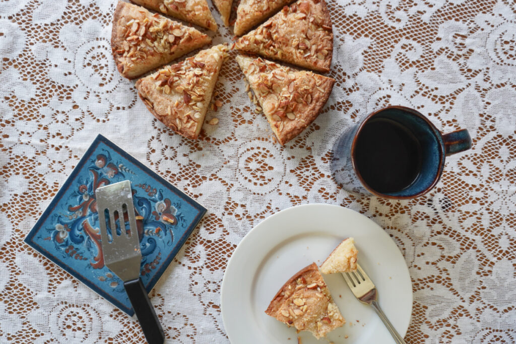 A table covered with a white lace tablecloth and set with slices of almond skillet cake and a mug of coffee