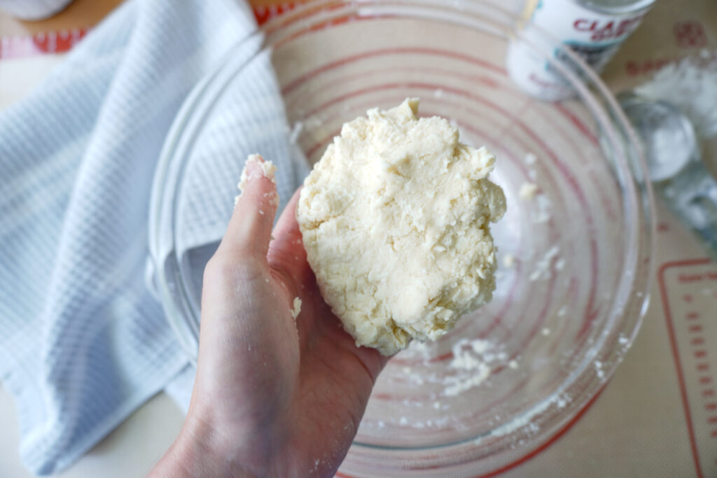 a hand holding a ball of pastry dough above a glass mixing bowl