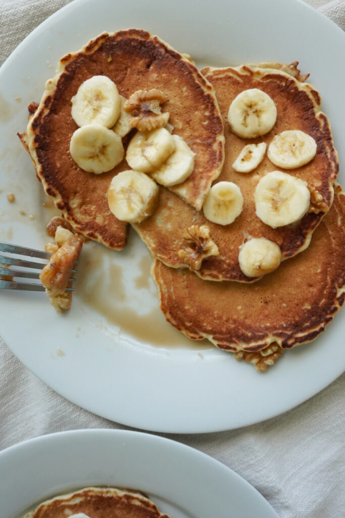 three banana nut pancakes spread out on a white plate topped with sliced bananas and walnuts and a bite taken out of one of the pancakes
