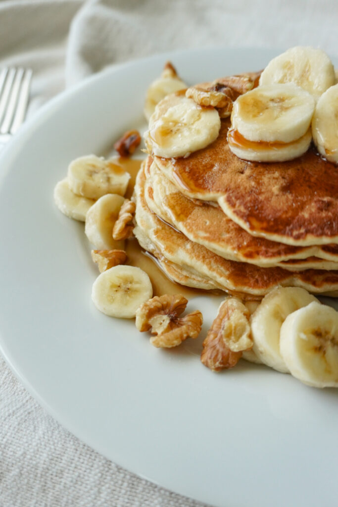 a view of the edges of a stack of pancakes on a white plate topped with bananas, walnuts, and maple syrup
