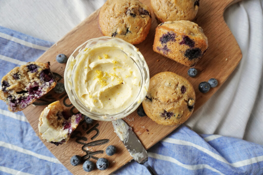 a wooden cutting board with blueberry muffins and a dish of whipped lemon honey butter on it