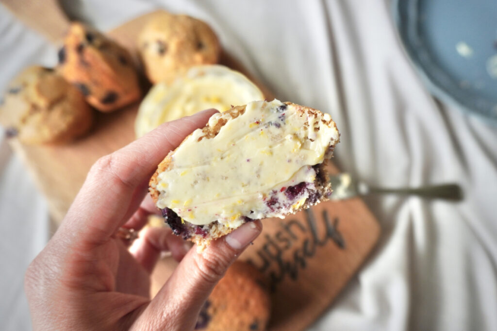a hand holding a halved blueberry muffin with whipped lemon honey butter spread on it