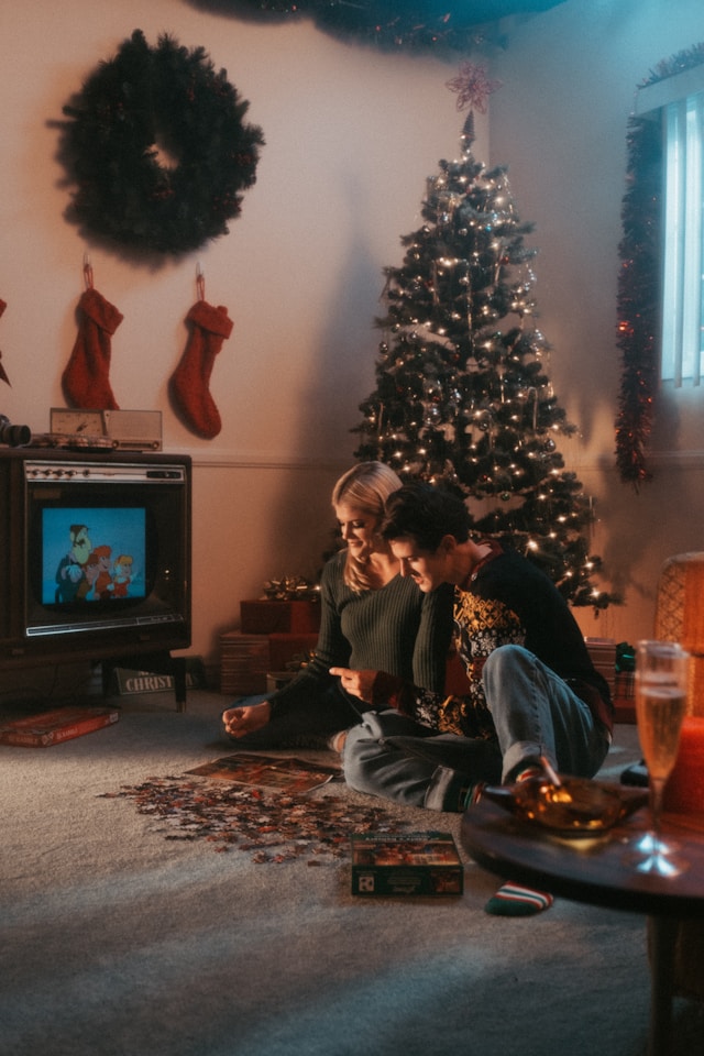 a couple sitting next to a lit christmas tree while doing a puzzle and watching cartoons